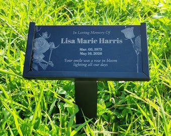 May include: A black memorial plaque with the inscription "In Loving Memory Of Lisa Marie Harris" and dates. The plaque features etched rose illustrations and is mounted on a black metal stake, set in a grassy area.