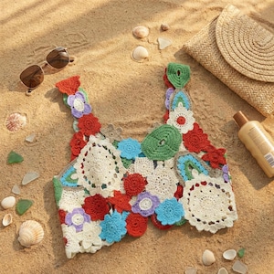 May include: A colorful, handmade crochet top with a floral design in red, blue, green, and white. The top is displayed on a sandy surface with sunglasses, a straw clutch, and seashells.