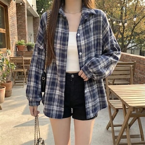 May include: A blue and white plaid button-down shirt worn over a white tank top and black shorts. The shirt has long sleeves and is unbuttoned. A black purse with a gold chain is visible.