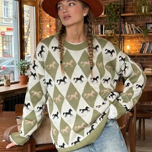 May include: A green and white argyle pattern jumper with black and tan horse silhouettes. The jumper has a crew neck and long sleeves with green trim. The person in the image is wearing a brown hat and blue jeans.