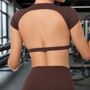 Puede incluir: Top deportivo marrón con mangas cortas y espalda abierta. El top se combina con leggings marrones a juego. La imagen está tomada en un gimnasio, mostrando ropa deportiva.