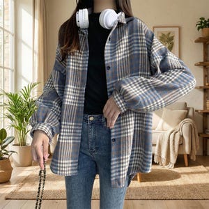 Puede incluir: Una camisa-chaqueta a cuadros azules, marrones y blancos con mangas largas y botones. La camisa-chaqueta se combina con vaqueros azules. Unos auriculares blancos cuelgan del cuello.