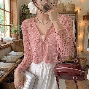 May include: A pale pink, open-weave cardigan with a V-neck and tie detail. The long-sleeved sweater has a scalloped hem and cuffs. A burgundy handbag with a top handle and zip closure is also visible.