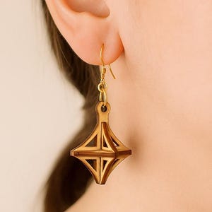 May include: A close-up of a gold earring with a geometric wooden pendant. The pendant is a diamond shape with an open, angular design. The earring is hanging from a gold hook, showcasing a unique, handcrafted jewelry piece.