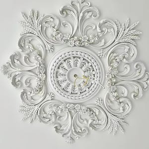May include: An elaborate white ceiling rose with detailed floral and scrollwork. The design includes a central circular feature and symmetrical patterns. The ceiling rose is mounted on a white ceiling, providing a decorative element.