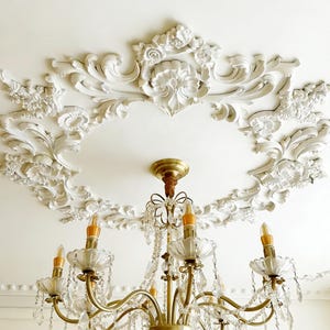 May include: A gold chandelier with crystal accents hangs from a white ceiling with ornate, floral plaster molding. The chandelier has six lights with amber-colored bulbs and dangling crystals, creating a classic, elegant aesthetic.