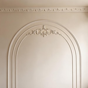 K&ouml;nnte beinhalten: Eine beige Wand mit einem bogenf&ouml;rmigen Stuckdesign und einem zentralen Zierornament aus Gips. Die Wand verf&uuml;gt oben &uuml;ber eine horizontale Deckenleiste mit wiederkehrendem Blumenmuster und unten &uuml;ber eine schwarze Sockelleiste.