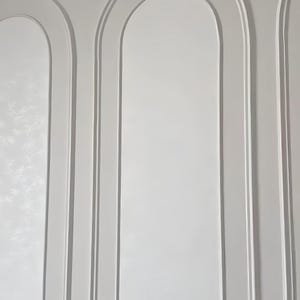 K&ouml;nnte beinhalten: Eine wei&szlig;e Wand mit drei vertikalen Bogenpaneelen, die durch erhabene Zierleisten definiert sind. Die Oberfl&auml;che ist glatt und matt, wobei jeder Bogen einen vertieften, abgerundeten oberen Bereich bildet, der der Wand Tiefe verleiht.