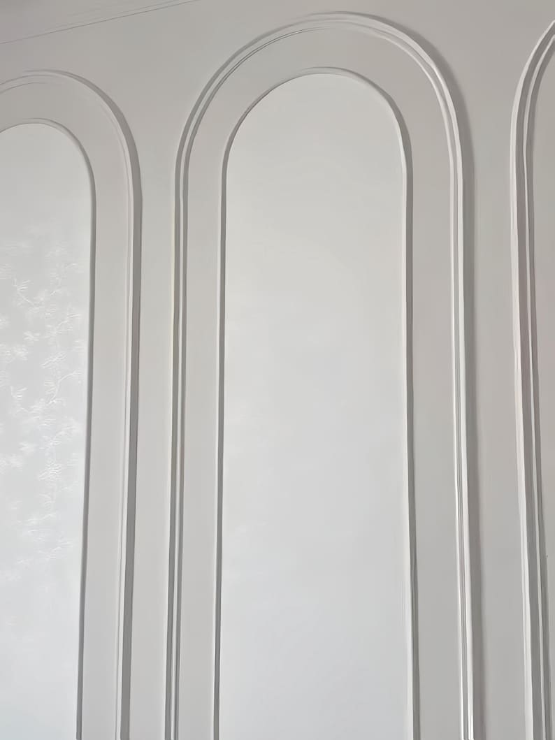 K&ouml;nnte beinhalten: Eine wei&szlig;e Wand mit drei vertikalen Bogenpaneelen, die durch erhabene Zierleisten definiert sind. Die Oberfl&auml;che ist glatt und matt, wobei jeder Bogen einen vertieften, abgerundeten oberen Bereich bildet, der der Wand Tiefe verleiht.