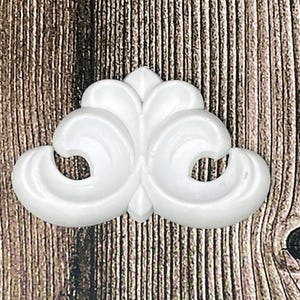 May include: A white, ornate decorative element with a symmetrical design. The piece features a central, pointed element flanked by two large, swirling scroll-like shapes. The background is a wood grain pattern.