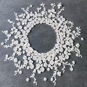 May include: A circular white floral design, possibly lace or fabric, arranged on a gray surface. The design features numerous small, detailed white roses and delicate, branching stems, creating a wreath-like appearance. The overall aesthetic is elegant and intricate.