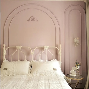 K&ouml;nnte beinhalten: Ein wei&szlig;es Metallbettgestell mit wei&szlig;er Steppbettw&auml;sche und zwei Kissen mit R&uuml;schen. Die Wand hinter dem Bett ist hellrosa gestrichen und mit bogenf&ouml;rmigen Zierleisten, einem dekorativen Wandelement und einer Wandleuchte versehen.