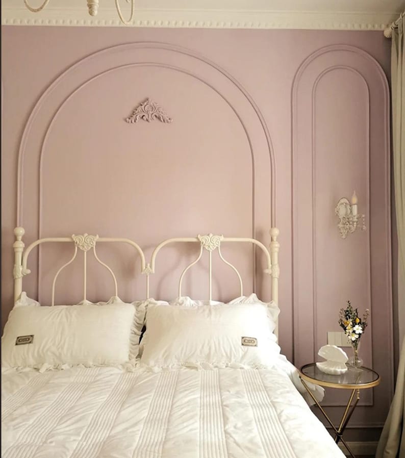 K&ouml;nnte beinhalten: Ein wei&szlig;es Metallbettgestell mit wei&szlig;er Steppbettw&auml;sche und zwei Kissen mit R&uuml;schen. Die Wand hinter dem Bett ist hellrosa gestrichen und mit bogenf&ouml;rmigen Zierleisten, einem dekorativen Wandelement und einer Wandleuchte versehen.