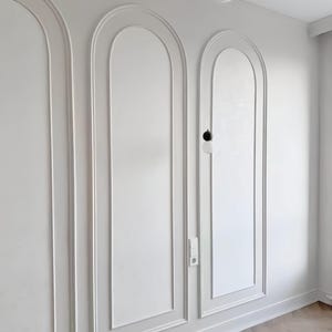 May include: White wall paneling with arched inset designs. The panels are framed with molding and create a decorative architectural feature. The room has a light wood floor and a white ceiling with cloud-shaped lighting.