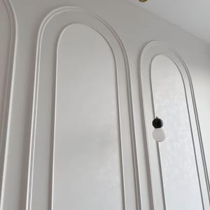 K&ouml;nnte beinhalten: Eine wei&szlig;e Wand mit drei gro&szlig;en, vertikalen Bogenleisten mit erhabenen R&auml;ndern. Eine einzelne Pendelleuchte h&auml;ngt von der Decke, bestehend aus einer kleinen schwarzen Kugel &uuml;ber einem gr&ouml;&szlig;eren wei&szlig;en Glaskugelschirm.