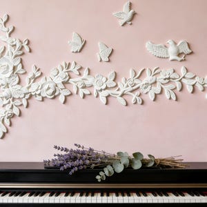Op de afbeelding: Een zwarte piano met witte toetsen staat onder witte bloemenwanddecoratie, vlinders en vogels tegen een roze muur. Een bosje lavendel en eucalyptus ligt bovenop de piano.