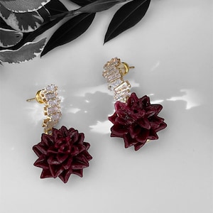 Könnte beinhalten: Ein Paar goldfarbene Ohrstecker. Jeder Ohrring hat eine Ansammlung klarer, rechteckiger Kristalle und einen tief burgunderfarbenen, blütenförmigen Anhänger. Die Ohrringe werden auf einer weißen Oberfläche mit dunkelgrünen und weißen Blättern im Hintergrund präsentiert.