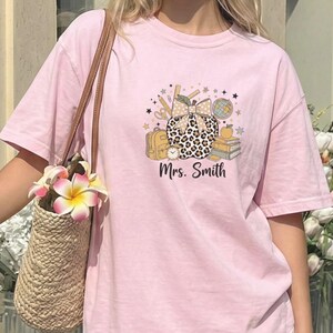 May include: A light pink short-sleeve t-shirt featuring a graphic of a leopard print apple with a bow, a backpack, books, and the text Mrs. Smith. The shirt is paired with a white pleated skirt and a woven straw bag with colorful flowers.