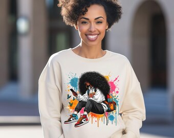 May include: Cream-colored sweatshirt featuring a colorful graphic of a person with large curly hair, sneakers, and a backpack. The graphic includes paint splatters and butterflies. The person in the graphic is holding a phone.