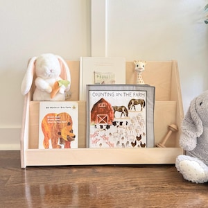 May include: A light-colored wooden bookshelf holding children's books titled Brown Bear, Brown Bear, What Do You See? and Counting on the Farm, accompanied by a white plush rabbit, a gray plush rabbit, a giraffe toy, and a small wooden rattle.
