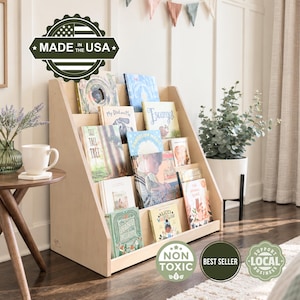 May include: A light-colored wooden bookshelf with four slanted tiers for displaying books. The unit features a triangular side profile and holds various children's books. A green circular badge with a flag icon reads MADE IN THE USA, NON TOXIC, BEST SELLER, and SUPPORT LOCAL BUSINESS.