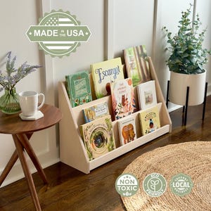 Op de afbeelding: Een lichtgekleurde houten boekenplank gevuld met kinderboeken. Een kleine ronde houten bijzettafel met een vaas met bloemen en een mok staat op de voorgrond. Een plant in een witte pot staat op de achtergrond. De afbeelding bevat een "Made in the USA"-logo.