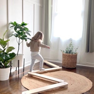 以下が含まれることがあります： 子供がジュートラグの上に置かれた木製のバランスビームでバランスをとっています。バランスビームは明るい色の木材でできており、ジグザグのデザインです。部屋には広葉樹の床、白い壁、シアーなカーテンのある大きな窓があります。鉢植えの植物が背景にあります。