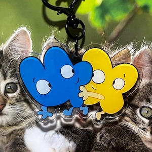 Puede incluir: Un llavero acrílico transparente con dos personajes de dibujos animados con forma del número cuatro. Uno es azul, el otro amarillo, ambos con ojos blancos. El llavero tiene un cierre negro y una bola blanca en la parte superior.