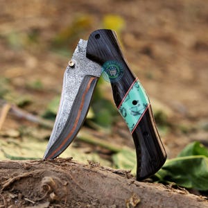 Peut inclure: Un couteau pliant avec une lame à motifs et un manche en bois foncé. Le manche présente une incrustation turquoise avec des accents rouges. La lame a un bord couleur cuivre. Le couteau est ouvert, montrant la lame.
