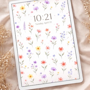 May include: A white tablet displays a floral wallpaper with watercolor-style flowers in yellow, purple, red, and blue. The time 10:21 and the date Tuesday, April 23 are shown on the screen. The tablet rests on a beige fabric.