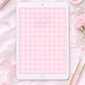 Może przedstawiać: Biały tablet wyświetla różowo-biały wzór w kratkę, z godziną 11:27 i datą wtorek, 23 kwietnia. Obraz zawiera spiralny notatnik, długopisy i kwiatowe akcenty, wszystko w miękkich różowych odcieniach.