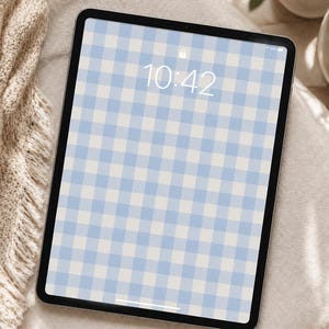 以下が含まれることがあります： 黒いタブレットの画面には、水色と白のギンガムチェック模様が表示されています。時刻は10:42です。タブレットはベージュ色の表面に置かれており、左側にはフリンジ付きの毛布の一部が見えています。