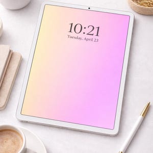 Puede incluir: Una tableta blanca muestra la hora 10:21 y la fecha martes 23 de abril, con un fondo degradado amarillo y rosa. Un bolígrafo blanco con detalles dorados, una taza de café y artículos de papelería también son visibles.