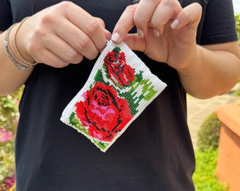 May include: A white beaded pouch featuring a vibrant design of red roses with green leaves. The pouch is rectangular and has a zipper closure. The floral pattern is detailed and eye-catching, perfect for carrying small items.