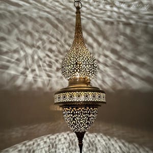 May include: A brass-colored, ornate pendant light fixture with a pierced metal design. The light is suspended from the ceiling and casts intricate shadow patterns on the wall. The fixture has a bulbous shape with a pointed bottom.