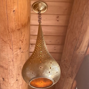 Puede incluir: Una lámpara colgante de latón con una pantalla en forma de lágrima. La pantalla tiene un diseño perforado, que permite que la luz se filtre. La lámpara está suspendida de una cadena y un soporte de techo, creando un ambiente cálido.
