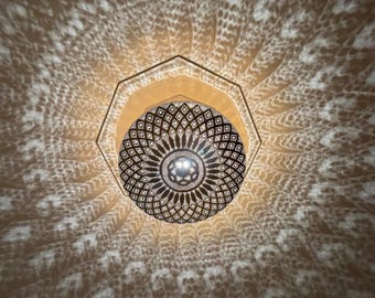 May include: A decorative, ornate pendant light with a circular, patterned design. The light casts intricate shadows on the surrounding surface, creating a beautiful display. The light fixture is suspended from a geometric frame.