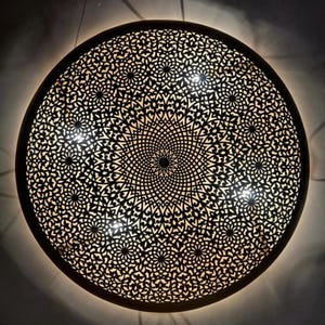Peut inclure: Un luminaire décoratif rond en métal noir avec un motif découpé complexe et symétrique. Le design présente des formes florales et géométriques, avec une lumière chaude émanant de derrière le métal. Le luminaire est fixé au mur.