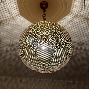 Può includere: Un lampadario sferico con un intricato design traforato. Il paralume in metallo è di un caldo colore oro e la luce proietta ombre decorative. L'apparecchio è appeso a una catena ed è montato su un soffitto bianco.
