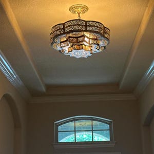 Pode incluir: Candelabro ornamentado de vários níveis com intrincados padrões recortados, suspenso de uma luminária de teto decorativa. O candelabro tem um tom dourado quente, projetando um brilho suave. O design é geométrico e apresenta uma forma de estrela.