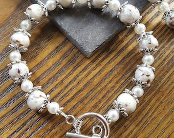 Op de afbeelding: Een witte en bruine kralen armband met een zilveren schuifgesp. De kralen zijn rond en hebben een marmerpatroon. De armband ligt op een houten oppervlak.