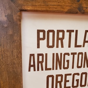 May include: A wooden framed sign with the words "PORTLAND, ARLINGTON, OREGON, CHICAGO" printed in brown lettering on a white background.