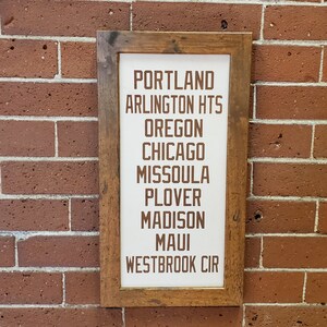 Puede incluir: Un letrero enmarcado en madera con fondo blanco y texto marrón. El texto dice "PORTLAND, ARLINGTON HTS, OREGON, CHICAGO, MISSOULA, PLOVER, MADISON, MAUI, WESTBROOK CIR".
