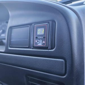 May include: Close-up of a black dashboard interior with a digital display. The display shows the number "01" and the word "HYDRA" with a graphic of a flame. The dashboard is made of textured black plastic.