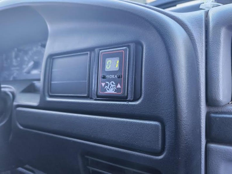 May include: Close-up of a black dashboard interior with a digital display. The display shows the number "01" and the word "HYDRA" with a graphic of a flame. The dashboard is made of textured black plastic.