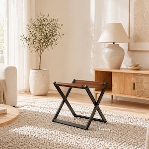 Könnte beinhalten: Ein Klapphocker mit braunem Ledersitz und schwarzem Metallrahmen. Der Hocker steht auf einem strukturierten Teppich in einem Wohnzimmer. Der Raum hat eine neutrale Farbgebung mit einem Sofa, einer Lampe und anderen dekorativen Gegenständen.