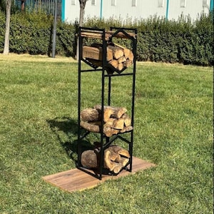 Op de afbeelding: Een zwart metalen houtrek met drie planken, elk met een stapel gezaagde blokken. Het rek staat op een rechthoekige houten basis, geplaatst op een grasveld. Het ontwerp is geometrisch en modern, geschikt voor buitengebruik.