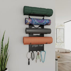 Op de afbeelding: Een zwarte metalen yogamattenrek aan een witte muur, met vier opgerolde yogamatten in groen, patroon, grijs en oranje. Onder de matten staat een bordje met "- yoga mat" met haken voor accessoires.
