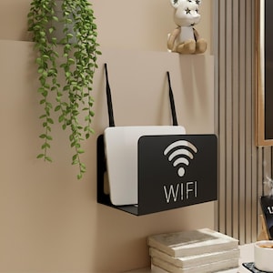 Puede incluir: Un enrutador WiFi blanco y negro con dos antenas está montado en una pared. El enrutador se sujeta en un soporte de metal negro con la palabra "WIFI" y un símbolo WiFi. Una planta verde cuelga de una maceta encima del enrutador.