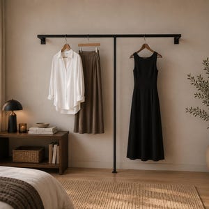 Pode incluir: Um cabideiro de parede em metal preto que sustenta uma camisa branca de manga comprida, uma saia midi castanha num cabide de madeira e um vestido midi preto sem mangas. O cabideiro está fixado numa parede sobre uma mesa de apoio em madeira.